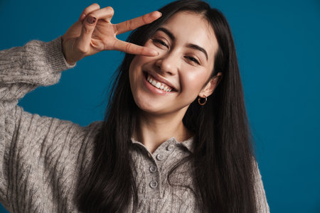 Happy beautiful asian girl smiling and showing peace sign isolated over blue backgroundの写真素材