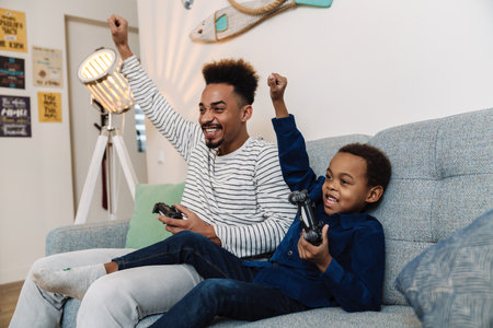 Excited african american father and son making winner gesture while playing video game at home