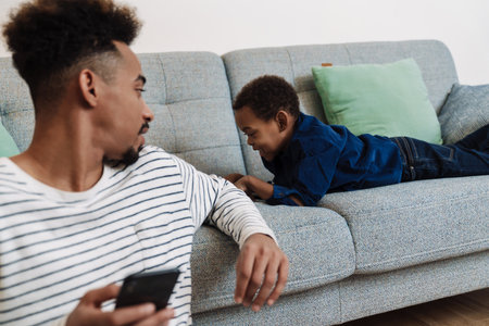 African american father and son using mobile phone while resting at homeの写真素材