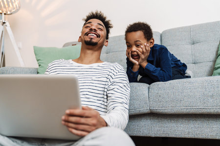 Happy african american father and son smiling and using laptop while sitting at home