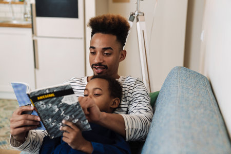 Focused african american father and son reading book while resting on couch at homeの写真素材
