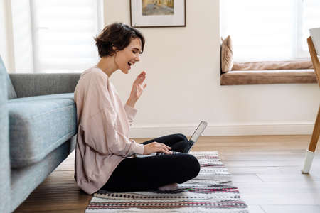 Excited beautiful girl waving hand while using laptop on floor at homeの写真素材