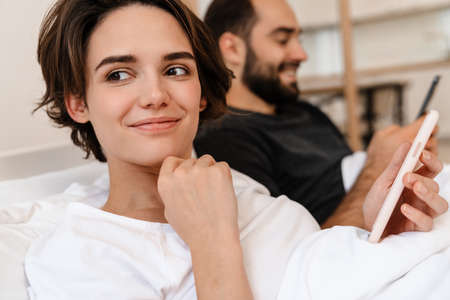 Happy young couple smiling and using mobile phones while lying in bed at home