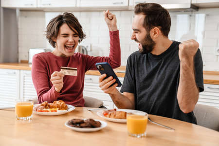 Excited couple making winner gesture using cellphone and credit card at home kitchenの写真素材