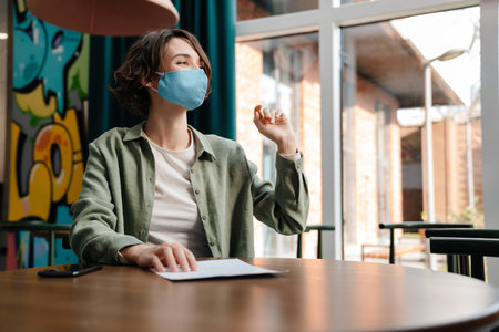 Happy girl in protective mask waving hand while sitting at cafe indoorsの写真素材