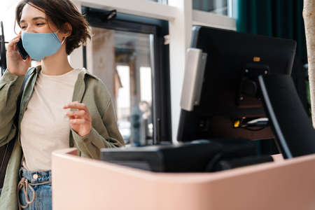 Happy girl in protective mask talking on cellphone while standing indoorsの写真素材