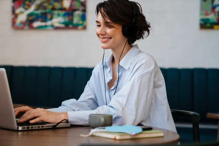 Happy nice girl wearing headphones working with laptop while sitting in cafe