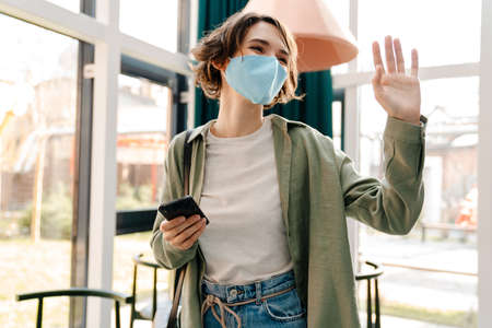 Happy girl in protective mask waving hand and using cellphone while standing in cafeの写真素材