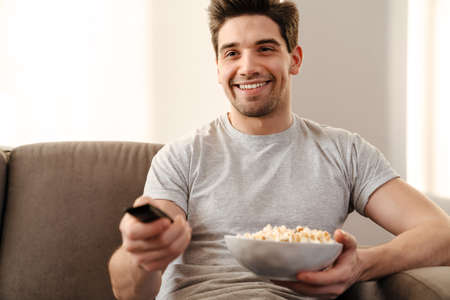 Happy man watching tv and eating popcorn at home on a couchの写真素材