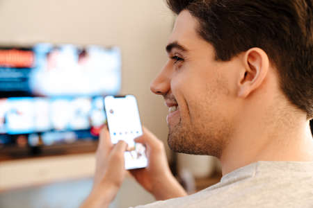 Caucasian smiling guy using mobile phone while watching TV at homeの写真素材