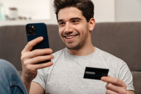 Happy brunette guy using mobile phone and credit card while sitting on floor at homeの写真素材