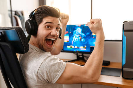 Excited guy in headphones making winner gesture while playing video game on computer at home