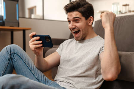 Delighted guy making winner gesture while playing video game on cellphone at homeの写真素材