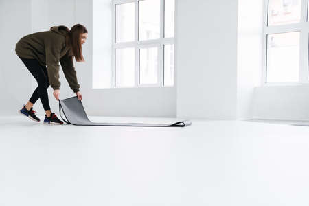 Athletic pleased sportswoman unrolling fitness mat before practice indoors