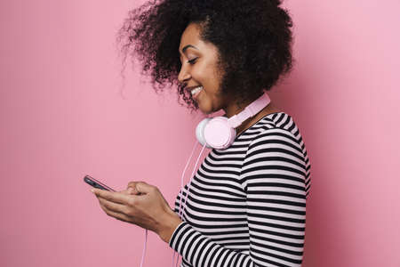 Joyful african american girl with headphones using cellphone isolated over pink background