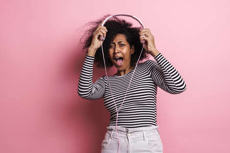 Joyful african american girl making fun with headphones isolated over pink background