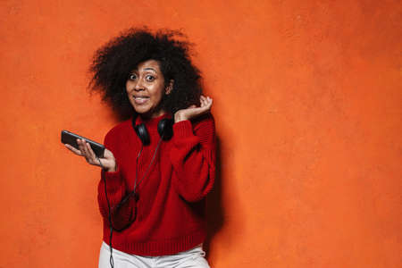 Smiling attractive casual young african woman with headphones standing isolated pver orange background, using mobile phoneの写真素材