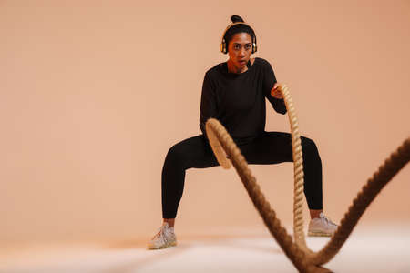 Athletic african american woman in headphones working out with battle ropes isolated over beige backgroundの写真素材