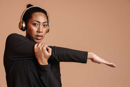 Focused african american woman in headphones doing exercise while working out isolated over beige...