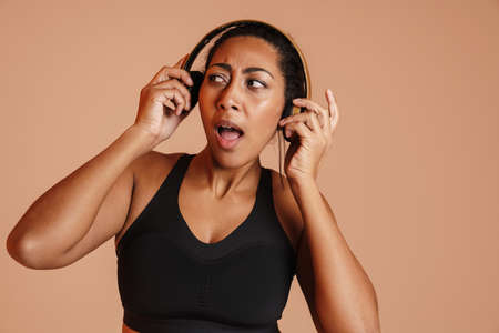Shocked african american sportswoman posing in wireless headphones isolated over beige background