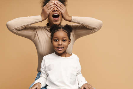 Happy black mother and daughter covering eyes while making fun isolated over beige backgroundの写真素材