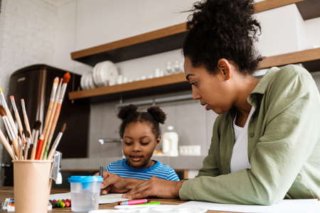 Black mother drawing with her daughter at table in home kitchen