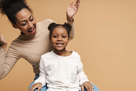 Happy black mother and daughter making fun while sitting isolated over beige backgroundの写真素材