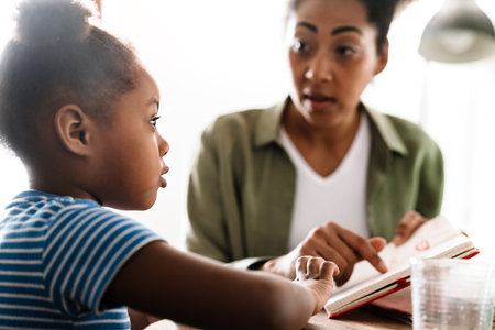 Black mother reading with her daughter at table in home kitchenの写真素材
