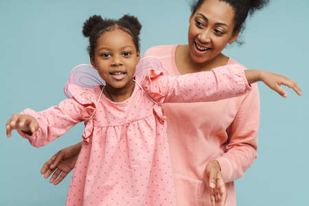 Happy black mother and daughter with butterfly wings making fun together isolated over blue...