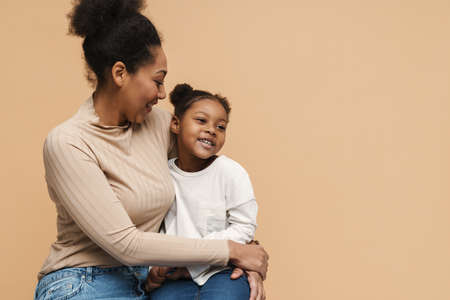 Smiling black mother and daughter hugging and sitting isolated over beige backgroundの写真素材