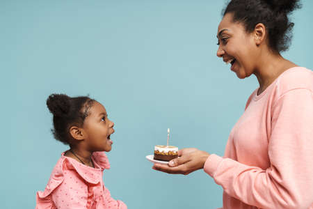 Happy black mother and daughter celebrating birthday with cake isolated over blue backgroundの写真素材
