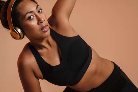 Focused african american woman in headphones doing exercise while working out isolated over beige...
