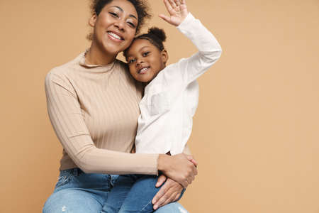 Happy black mother and daughter hugging and waving hand isolated over beige backgroundの写真素材