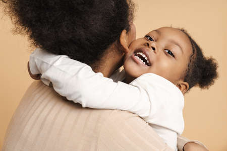 Happy black mother and daughter hugging and smiling isolated over beige background
