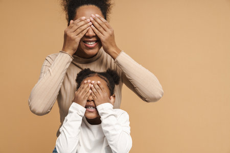 African mother and daughter smiling and covering their eyes isolated over beige wallの写真素材