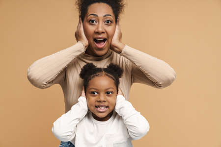 African mother and daughter posing and covering their ears isolated over beige wallの写真素材