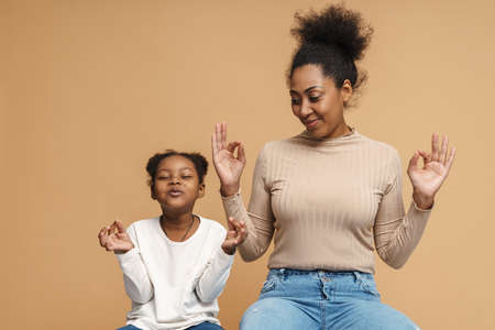 Smiling black mother and daughter gesturing while meditating together isolated over beige backgroundの写真素材