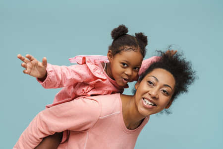 Joyful black mother and daughter with butterfly wings doing piggyback ride isolated over blue backgroundの写真素材