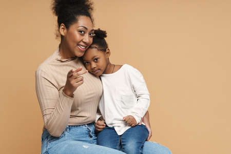 Happy black mother and daughter hugging while pointing finger at camera isolated over beige backgroundの写真素材