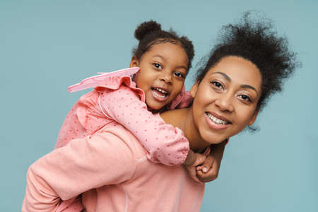 Joyful black mother and daughter with butterfly wings doing piggyback ride isolated over blue backgroundの写真素材