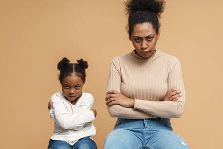 Displeased black mother and daughter frowning while sitting with arms crossed isolated over beige backgroundの写真素材