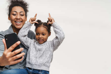 African mother and daughter making fun while using cellphone isolated over white wall
