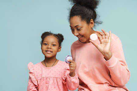 Happy black mother and daughter posing with macaroons isolated over blue backgroundの写真素材