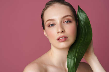 Beauty portrait of an attractive young woman posing with green leaf isolated over pink backgroundの写真素材