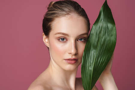 Beauty portrait of an attractive young woman posing with green leaf isolated over pink backgroundの写真素材