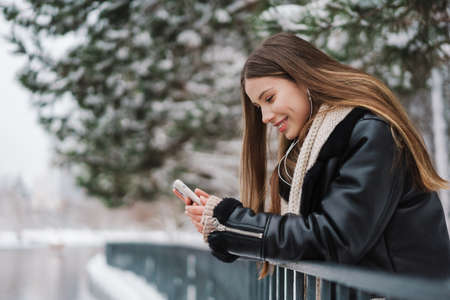 Happy girl in earphones using cellphone and smiling while walking in winter parkの写真素材