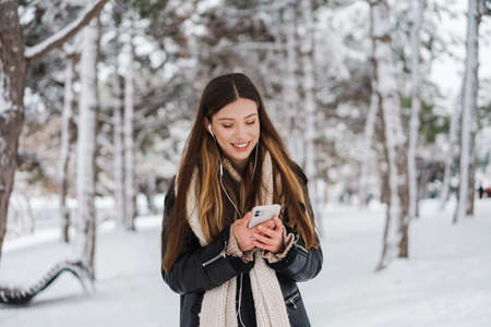 Happy girl in earphones using cellphone and smiling while walking in winter forestの写真素材