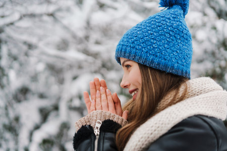Happy beautiful girl in knit hat warming her hands while walking in winter forest