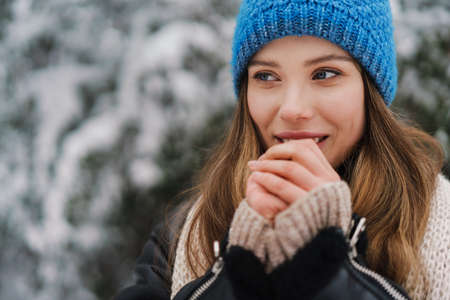 Happy beautiful girl in knit hat warming her hands while walking in winter forest