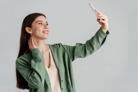 Happy caucasian girl smiling and taking selfie on mobile phone isolated over white backgroundの写真素材